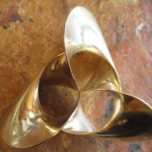 A rounded, shiny brass surface with threefold symmetry comprising a twisted band, against a reddish tile background. One edge of the band is a circle; the other edge wraps three full turns about the circle in a normal plane as that plane sweeps around the circle.