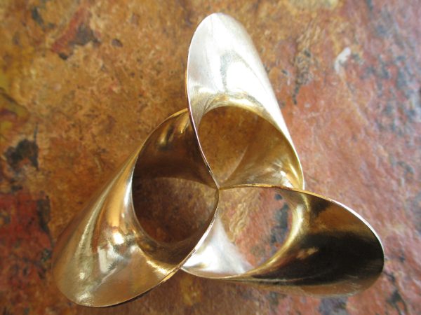 A rounded, shiny brass surface with threefold symmetry comprising a twisted band, against a reddish tile background. One edge of the band is a circle; the other edge wraps three full turns about the circle in a normal plane as that plane sweeps around the circle.
